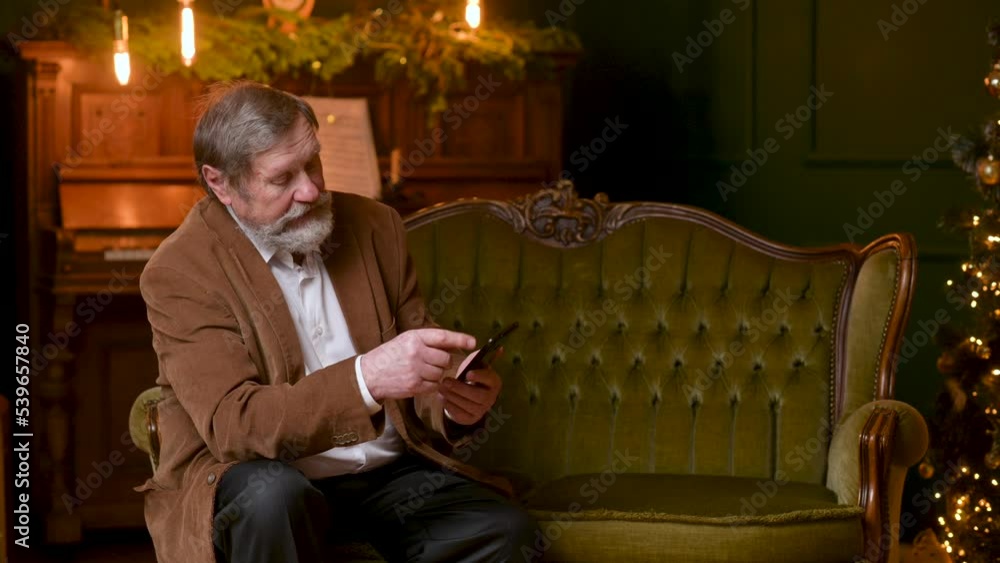 Elderly man sitting on the sofa in the room against the background of the Christmas tree and online video chatting. Christmas and New Year celebration Elderly man sitting on the sofa in the room against the background of the Christmas tree and online video chatting. Christmas and New Year celebration