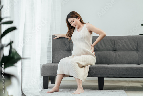 Full body of a beautiful pregnant asian woman is sitting on a couch in the living room, having a back pain as she carries the baby in her tummy. Pregnant problem.