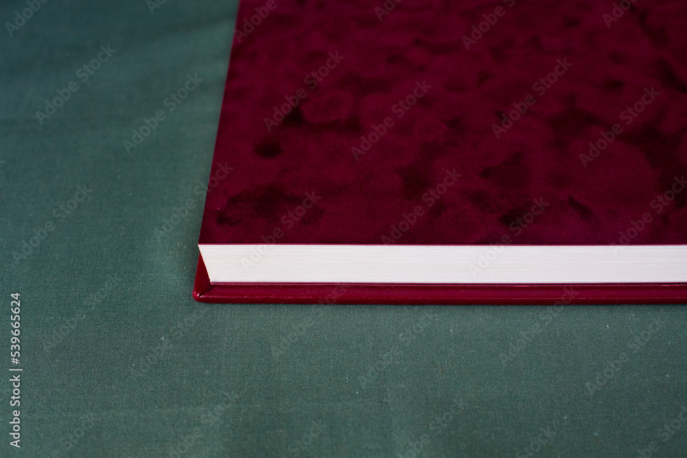 The velvet burgundy flyleaf of an expensive Premium photo book, with a white leather cover, lies open on a gray-green fabric in the room. The velvet burgundy flyleaf of an expensive Premium photo book, with a white leather cover, lies open on a gray-green fabric in the room.