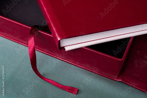A large stylish box and an expensive premium burgundy photobook with a leather cover and white pages lie on a gray table indoors.