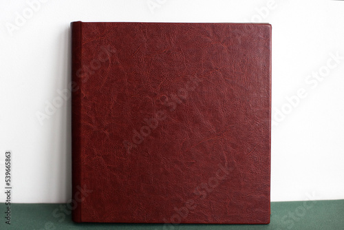 A large expensive premium photo book in a brown leather cover stands against a white wall on a green cloth indoors.