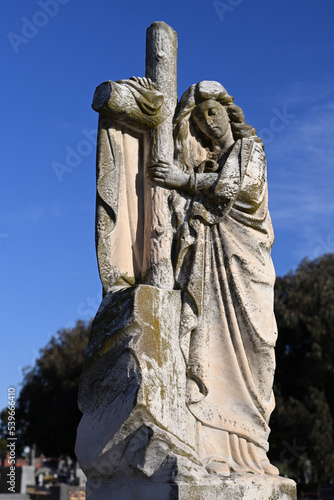 Worn and mossy stone sculpture of a woman wearing a robe while leaning on a crucifix in a cemetery  with bright blue sky in the background
