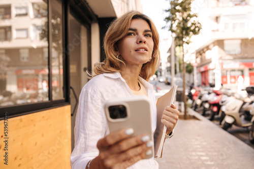 Modern young caucasian girl with phone and notebooks in her hands looks towards street during day. Blonde wears white shirt in spring. technology concept