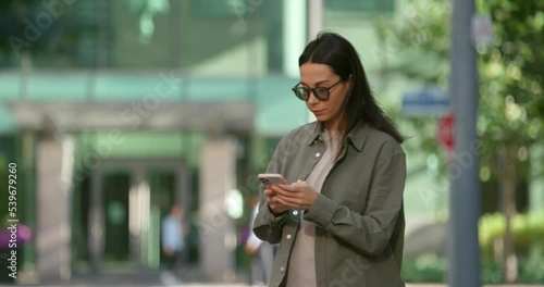 Attractive caucasian young woman with dark hair standing on the street, chatting and raising hand calling taxi, dressed in smart casual cloth 