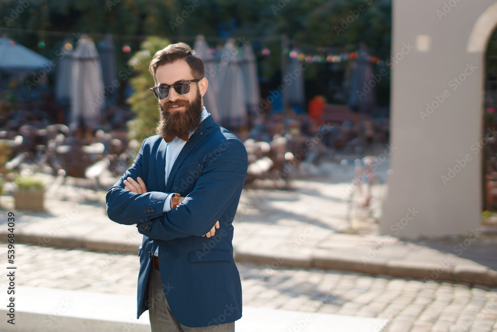 Stylish man with beard on the sunny street with crossed hands on his chest