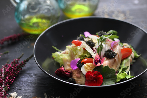 Salad with vegetables, fresh fish, caviar and quail egg, decorated with edible flowers. Salad in a black bowl, close up.