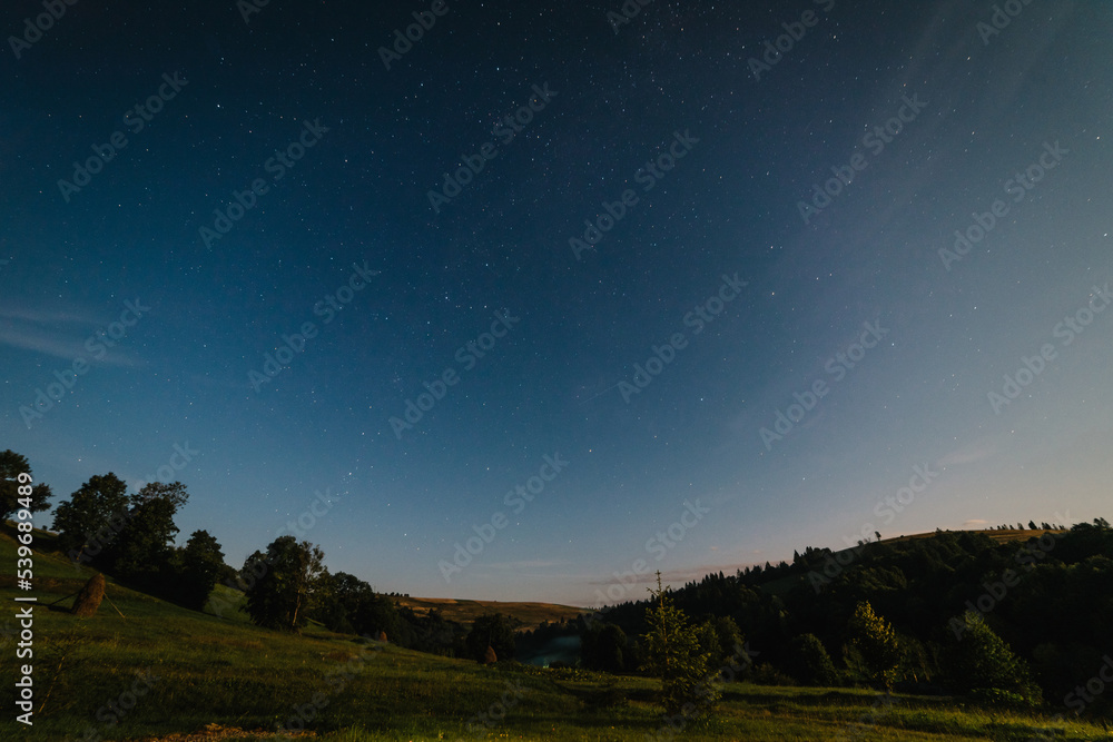 Night sky over the beautiful mountains, forest and green meadows in summer. Colorful landscape with bright starry sky with moonlight, constellation. Galaxy. Nature and space. Stars at night.