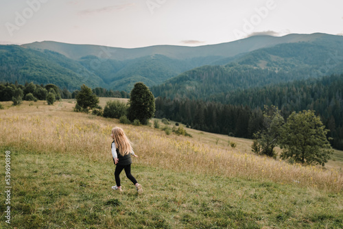 Child in autumn mountains. Girl is running on the grass in the field and enjoying nature. Little hiker and traveler walk on top of foggy mountain. World Tourism Day. Active outdoor games. Back view. © Serhii