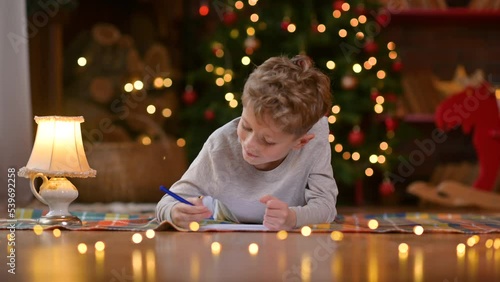 Portrait of a little boy lies on the floor and writes a letter to Santa Claus against the background of a festive Christmas tree. holidays and celebrations concept