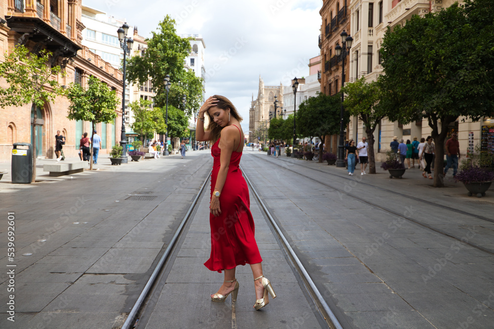 beautiful young woman in a red silk dress stands between the tram tracks on an avenue in seville. The model poses for the photo shoot. Fashion and beauty concept.