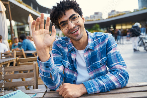 Handsome smiling student calling to teacher, online lesson concept. Middle eastern hipster man talking waving hand having video conference sitting outdoors