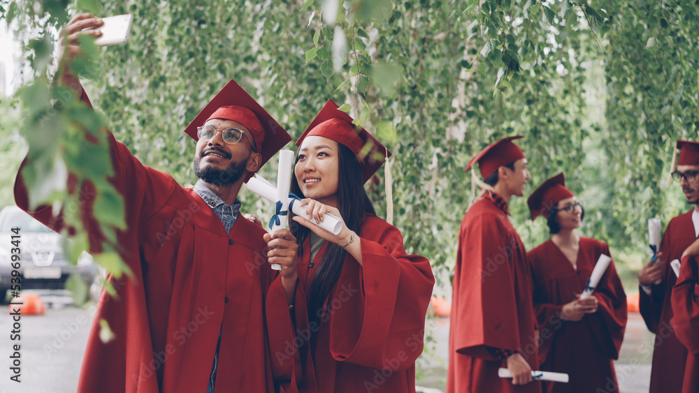 Obraz premium Two students are taking selfie with graduation diplomas wearing mortarboards and gowns, guy is holding smartphone and taking picture, girl is posing.