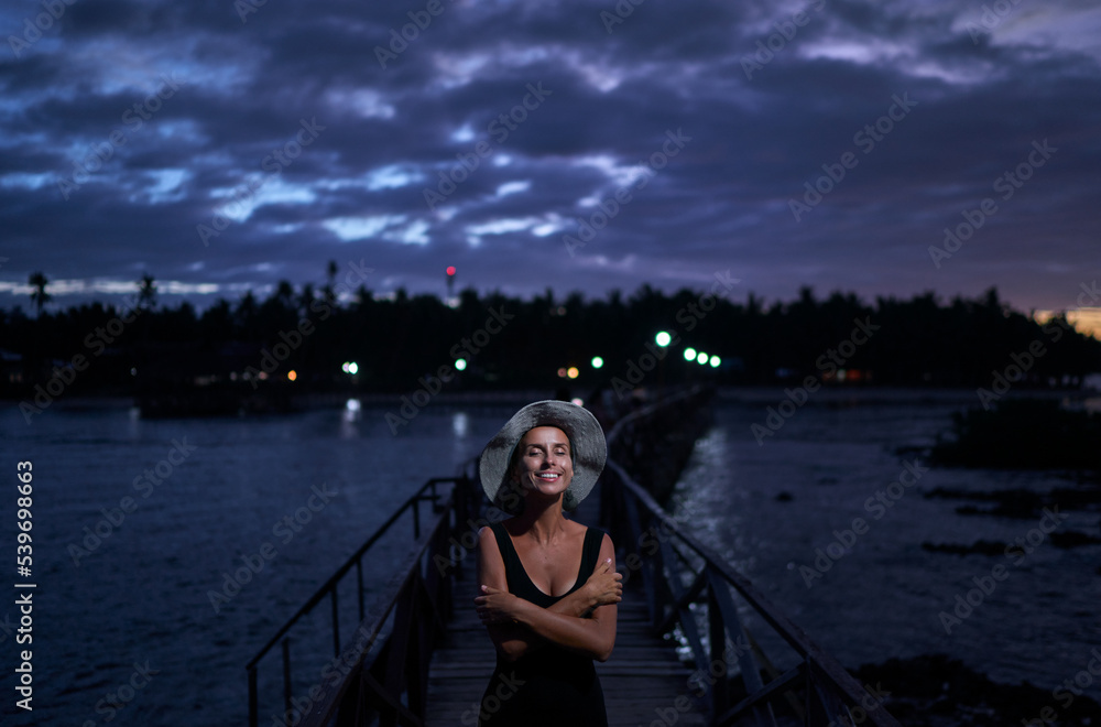 Obraz premium Vacation on tropical island. Young woman in hat enjoying sea view from wooden bridge terrace at night time, Siargao Philippines.