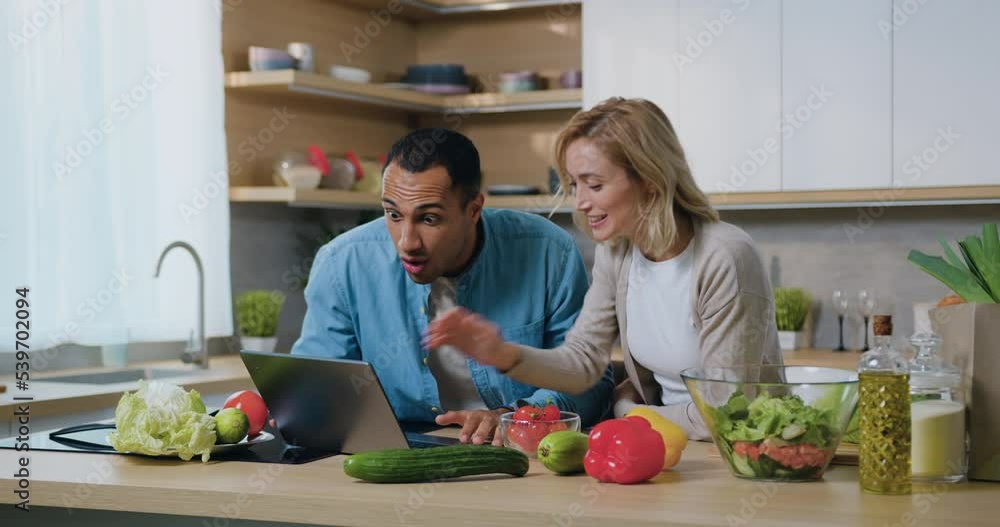 Multiethnic couple sit at kitchen with laptop check e-mail making yes gesture feel happy celebrate victory giving high five. African American man and woman readind good news on laptop. Multiethnic couple sit at kitchen with laptop check e-mail making yes gesture feel happy celebrate victory giving high five. African American man and woman readind good news on laptop.