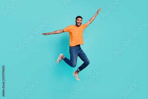 Full length photo of impressed funny guy wear orange t-shirt jumping high arms sides isolated teal color background