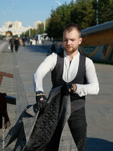 Stylish young man with a beard in classic clothes puts on a coat in a city park. Style, fashion, gentleman, clothing. Blue sky, autumn. photo