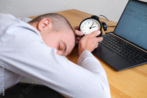 Person holds alarm clock in his hands and sleeps on table at the workplace next to a laptop