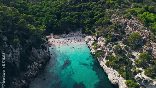 Aerial drone view of people having a good time by the beach with turquoise water. Tourists enjoy Cala Macarelleta and Macarella on the Balearic Islands, Spain, swimming and sailing yachts and boats. photo
