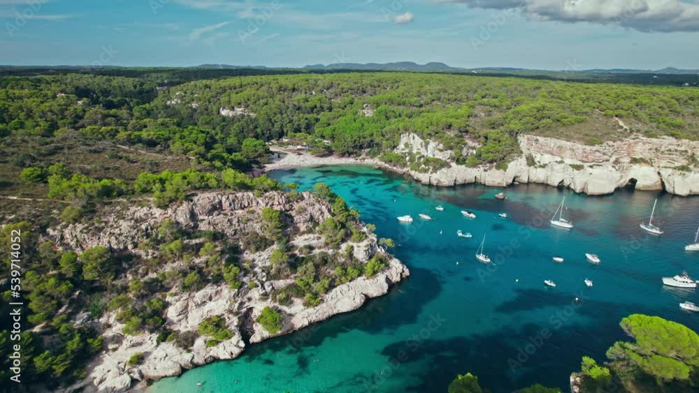 Aerial drone view of people having a good time by the beach with turquoise water. Tourists enjoy Cala Macarelleta and Macarella on the Balearic Islands, Spain, swimming and sailing yachts and boats. Aerial drone view of people having a good time by the beach with turquoise water. Tourists enjoy Cala Macarelleta and Macarella on the Balearic Islands, Spain, swimming and sailing yachts and boats.