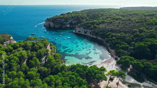 Aerial drone view of people having a good time by the beach with turquoise water. Tourists enjoy Cala Macarelleta and Macarella on the Balearic Islands, Spain, swimming and sailing yachts and boats.