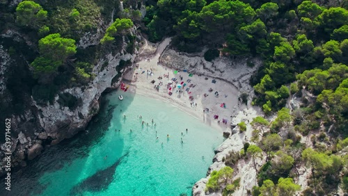 Aerial drone view of people having a good time by the beach with turquoise water. Tourists enjoy Cala Macarelleta and Macarella on the Balearic Islands, Spain, swimming and sailing yachts and boats. photo