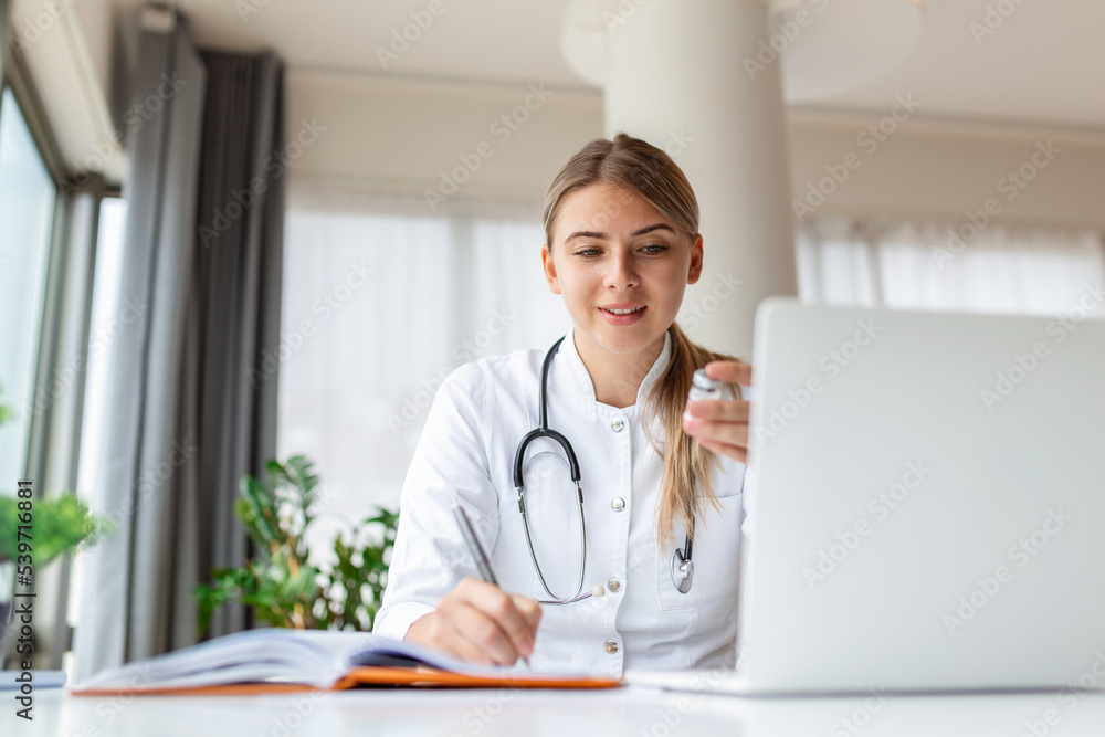 Smiling professional female doctor wearing uniform taking notes in medical journal, filling documents, patient illness history, looking at laptop screen, student watching webinar Smiling professional female doctor wearing uniform taking notes in medical journal, filling documents, patient illness history, looking at laptop screen, student watching webinar