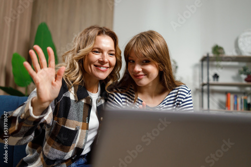 Happy mature woman waves her hand in greeting during a video call while sitting with her daughter in front of a laptop on the sofa in the living room