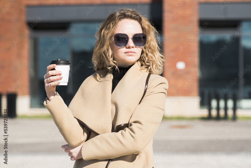 A young Caucasian girl in a coat drinks coffee and thinks while walking in the city in autumn. A young Caucasian girl in a coat drinks coffee and thinks while walking in the city in autumn.