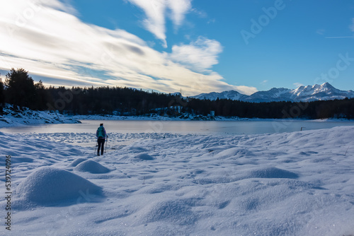 Woman with hiking backpack on field of deep crystal snow with scenic view of frozen alpine lake Forstsee, Techelsberg, Carinthia (Kaernten), Austria, Europe. Snow capped Karawanks, Mittagskogel (Kepa)