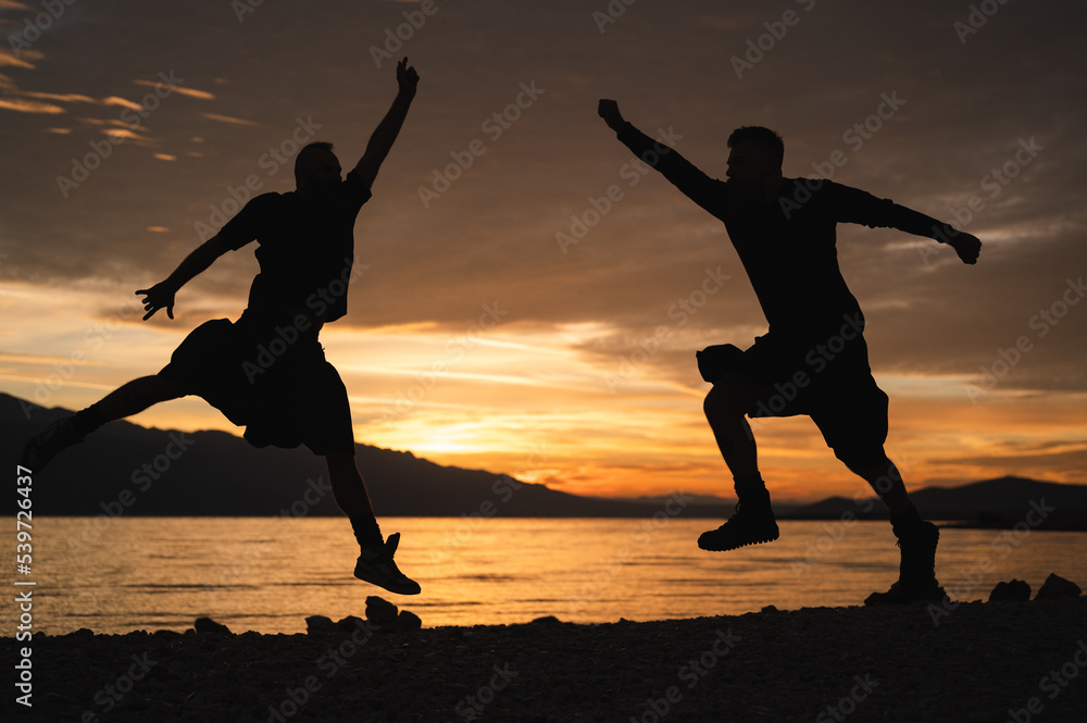 Gay couple in kilts jumping on the beach in sunrise. Silhouette of homosexual partners having early morning fun by seaside.