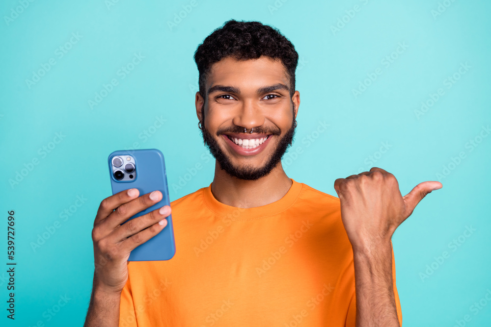 Photo of positive good mood guy wear orange t-shirt holding modern gadget thumb empty space isolated teal color background Photo of positive good mood guy wear orange t-shirt holding modern gadget thumb empty space isolated teal color background