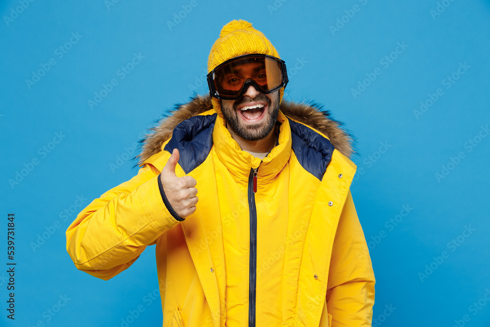 Young shocked fun skier man 20s wear yellow puffer down jacket with fur hood googles mask show thumb up isolated on plain blue color background studio. People winter lifestyle sport activity concept. Young shocked fun skier man 20s wear yellow puffer down jacket with fur hood googles mask show thumb up isolated on plain blue color background studio. People winter lifestyle sport activity concept.