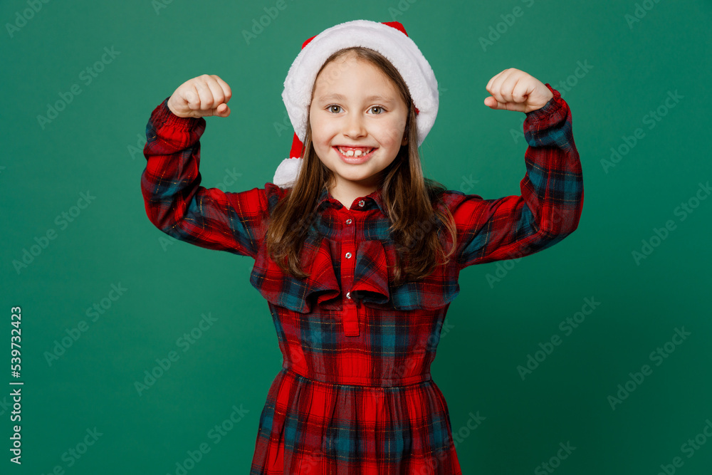 Merry little child kid girl 7 years old wear red dress Christmas hat posing showing biceps muscles on hand demonstrating strength power isolated on plain green background. Happy New Year 2023 concept. Merry little child kid girl 7 years old wear red dress Christmas hat posing showing biceps muscles on hand demonstrating strength power isolated on plain green background. Happy New Year 2023 concept.
