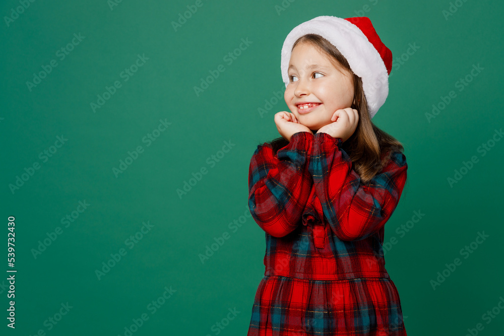 Merry smiling happy little child kid girl 6-7 years old wear red dress Christmas hat posing hold face look aside on workspace area isolated on plain dark green background. Happy New Year 2023 concept. Merry smiling happy little child kid girl 6-7 years old wear red dress Christmas hat posing hold face look aside on workspace area isolated on plain dark green background. Happy New Year 2023 concept.