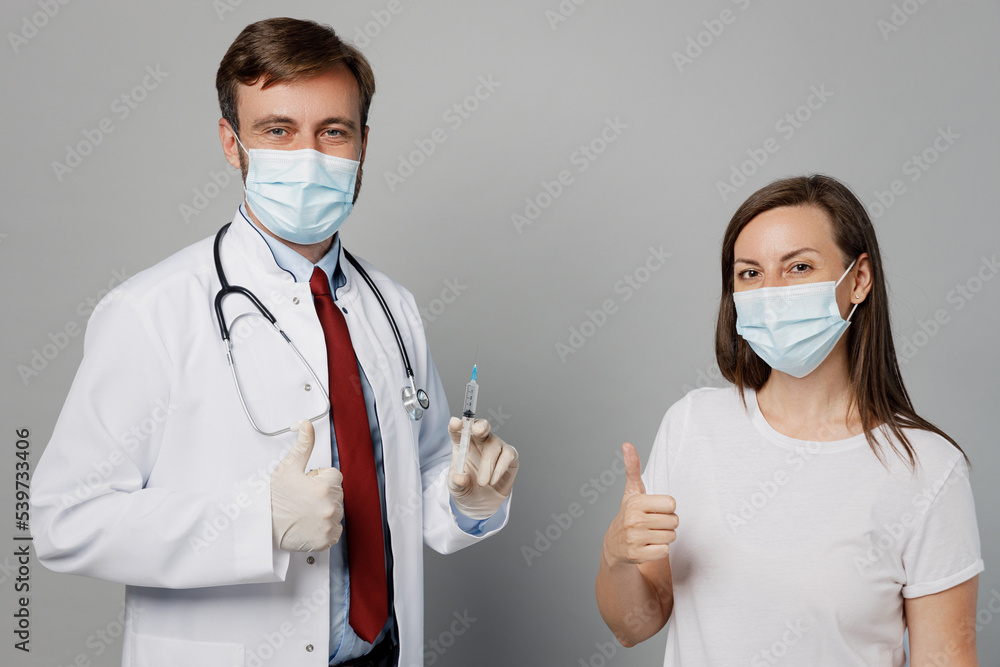 Male doctor man wear white medical gown suit mask gloves work in hospital hold syringe going to do injection of vaccine for girl patient isolated on plain grey background. Healthcare medicine concept. Male doctor man wear white medical gown suit mask gloves work in hospital hold syringe going to do injection of vaccine for girl patient isolated on plain grey background. Healthcare medicine concept.
