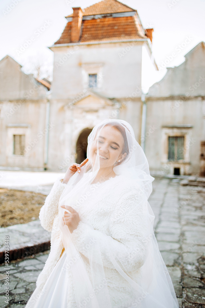 Lovely red-haired bride in a satin white dress with a veil poses in the courtyard of an ancient castle.
