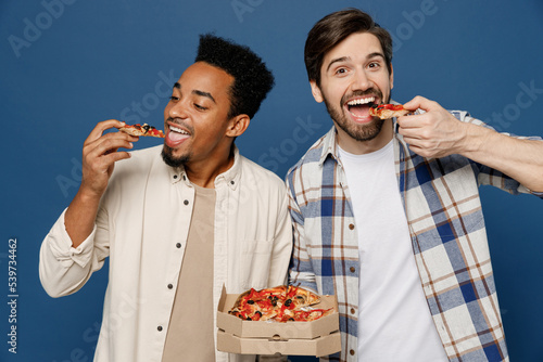 Young two friend men wear white casual shirts together holding italian pizza in cardboard flatbox eat biting slices look camera isolated plain dark royal navy blue background People lifestyle concept photo