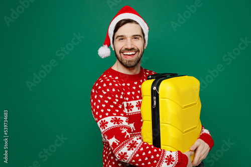 Traveler man wear knitted sweater Santa hat hold valise isolated on plain dark green background. Tourist travel abroad in free spare time rest getaway. Air flight trip journey concept New Year 2023. #539734683