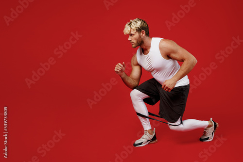 Full size side view young sportsman man wearing white clothes spend time in home gym train do squats lunges exercise with rubber band isolated on plain red background. Workout sport fit body concept. photo