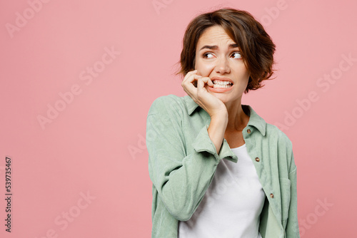 Fotomural Young woman 20s she wear green shirt white t-shirt look aside on workspace area biting nails fingers say oops isolated on plain pastel light pink background studio portrait