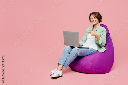 Full body young IT woman in green shirt white t-shirt sit in bag chair hold use work point finger on laptop pc computer isolated on plain pastel light pink background studio People lifestyle concept
