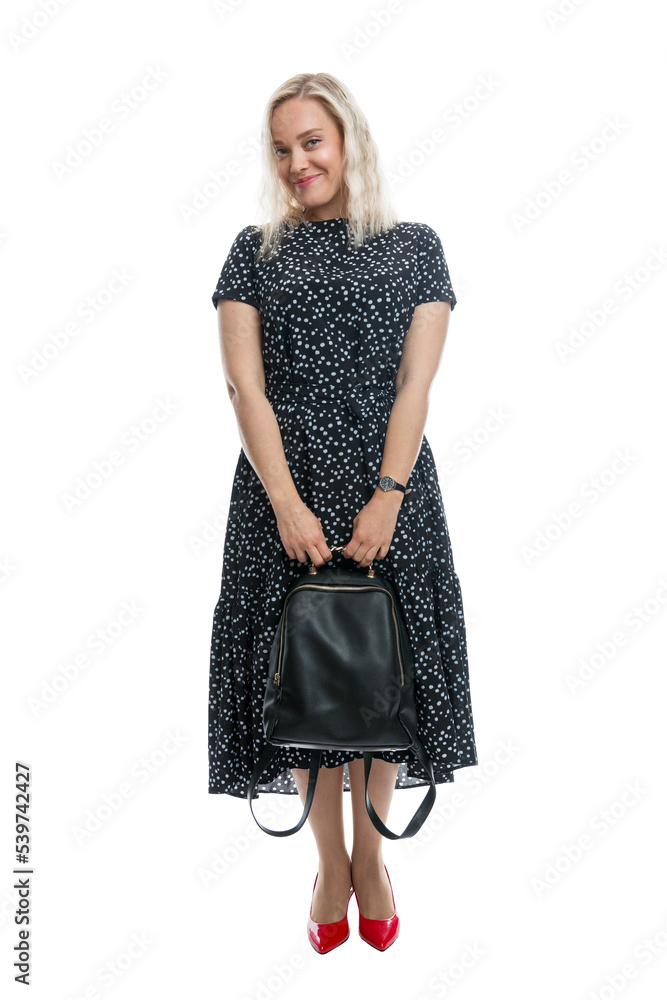 Beautiful smiling girl with a backpack in her hands. Blonde in a black romantic dress and red shoes. Joy and positivity. Isolated on a white background. Vertical. Beautiful smiling girl with a backpack in her hands. Blonde in a black romantic dress and red shoes. Joy and positivity. Isolated on a white background. Vertical.