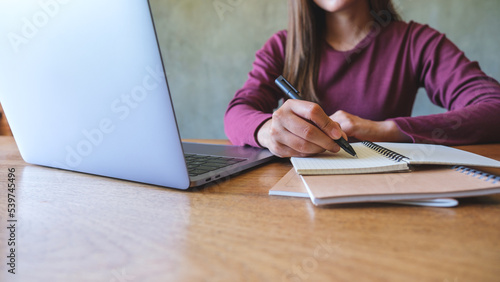 Closeup image of a young woman writing on a notebook while working on laptop computer