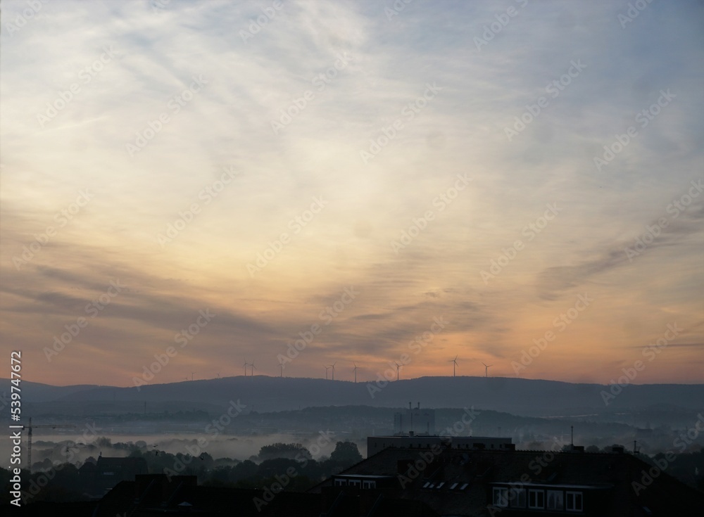 Sonnenaufgang über Windmühlen, Wald und Häuserdächern in Stadt am frühen Morgen bei Nebel im Herbst
