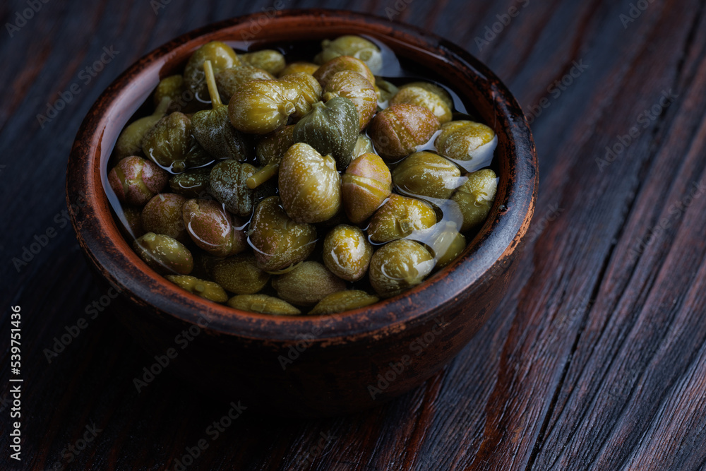 An earthenware bowl filled to the brim with capers stands on a wooden table. An earthenware bowl filled to the brim with capers stands on a wooden table.