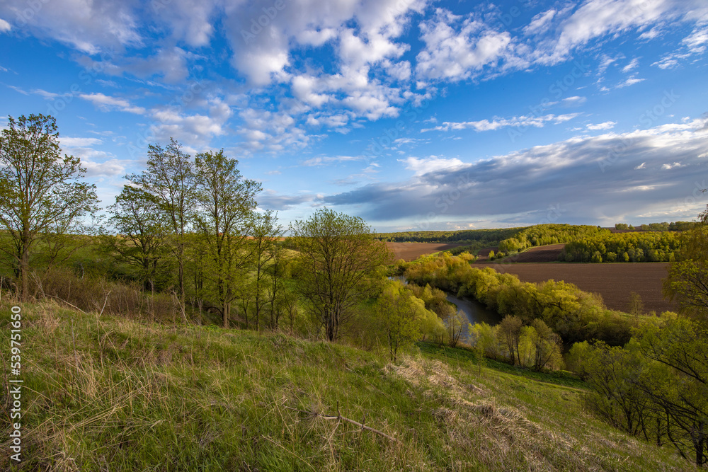 View from the hill to plowed farmland. Landscape with a river, arable land and green trees. View from the hill to plowed farmland. Landscape with a river, arable land and green trees.