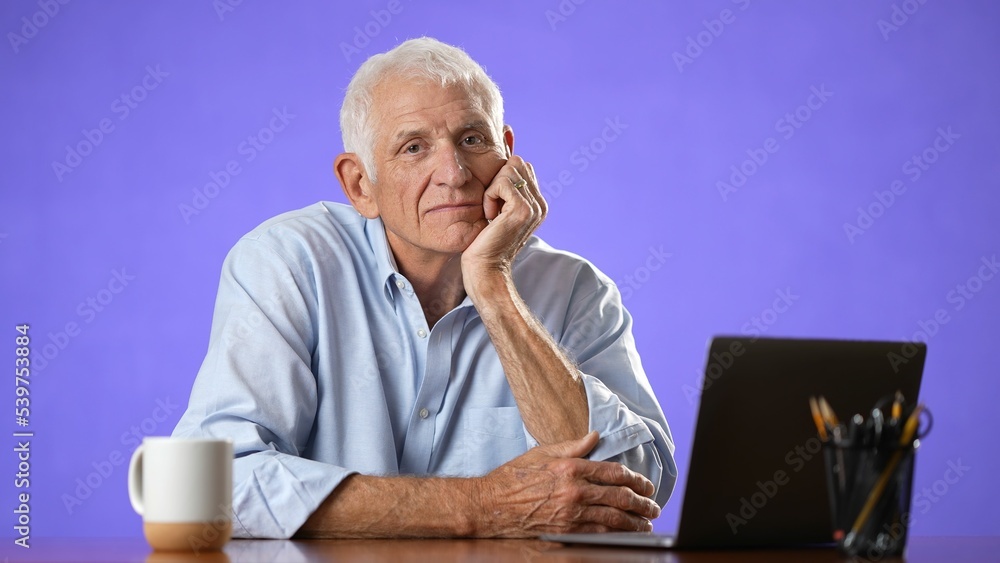 Portrait of happy older elderly man sitting at desk with laptop in home office. Confident 70s businessman with gray hair smiling isolated on solid purple background. Portrait of happy older elderly man sitting at desk with laptop in home office. Confident 70s businessman with gray hair smiling isolated on solid purple background.