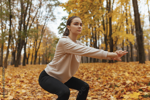Fit focused sportswoman doing squat exercise while working out in autumn park 