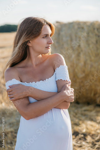 beautiful pregnant brunette in a white dress near the haystacks in the field at sunset. High quality photo