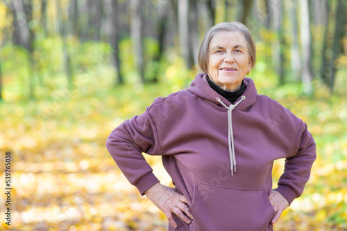 Fototapeta Naklejka Na Ścianę i Meble -  Portrait of a smiling elderly woman in a purple hoodie against the backdrop of an autumn park. Keeps hands on waist. Warm-up outdoors.